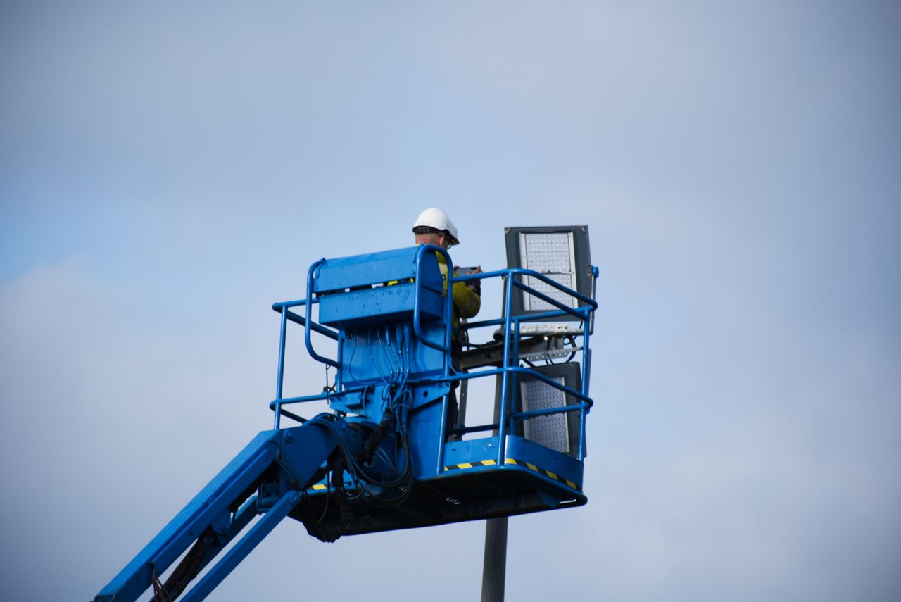 An industrial worker wearing a hard hat operating a lift platform for maintenance tasks in an outdoor setting.
